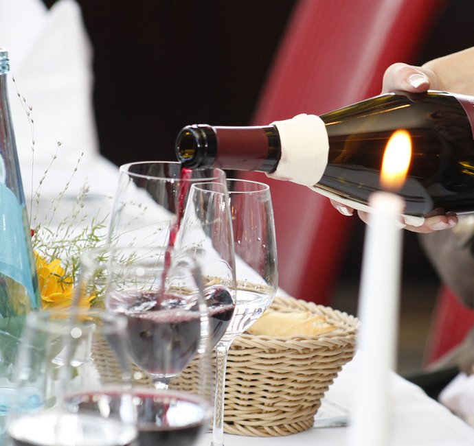 Ein Glas auf einem Tisch vor einem Brotkorb. In das Glas wird von rechts aus einer Flasche Rotwein gegossen. 
