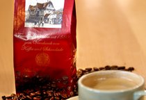 Eine Tüte steht in losen Kaffeebohnen neben einer Tasse. Auf der Tüte ist der Schriftzug "SchillerGarten. Espresso. Familienkonzession seit 1880 zum Verschank von Kaffee und Schokolade" zu sehen.