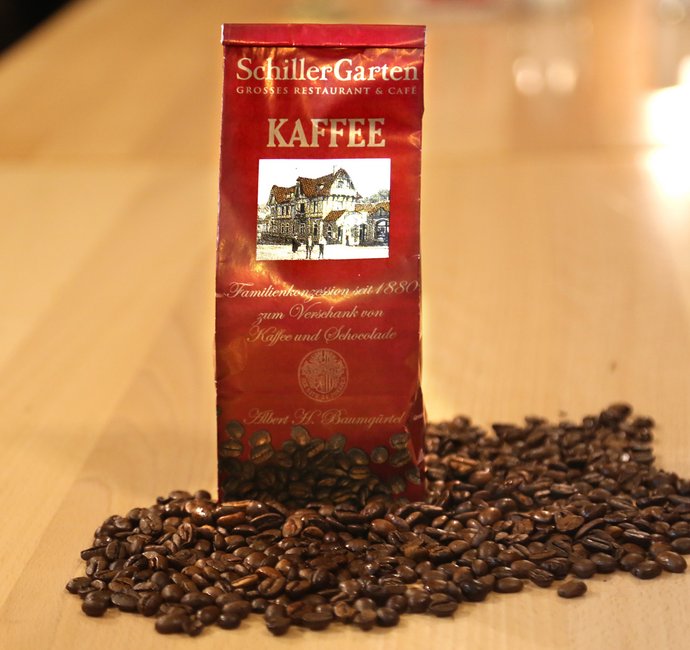 Eine Tüte steht in losen Kaffeebohnen. Auf der Tüte ist der Schriftzug "SchillerGarten. Kaffee. Familienkonzession seit 1880 zum Verschank von Kaffee und Schokolade" zu sehen.