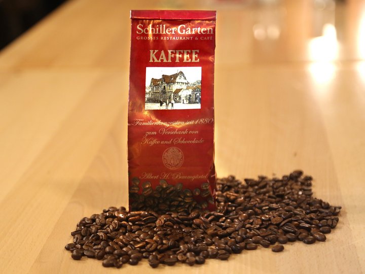 Eine Tüte steht in losen Kaffeebohnen. Auf der Tüte ist der Schriftzug "SchillerGarten. Kaffee. Familienkonzession seit 1880 zum Verschank von Kaffee und Schokolade" zu sehen.