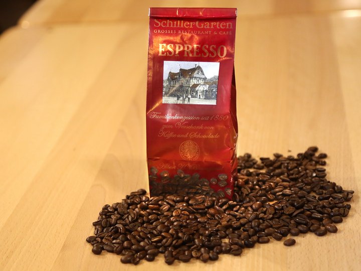 Eine Tüte steht in losen Kaffeebohnen. Auf der Tüte ist der Schriftzug "SchillerGarten. Espresso. Familienkonzession seit 1880 zum Verschank von Kaffee und Schokolade" zu sehen.