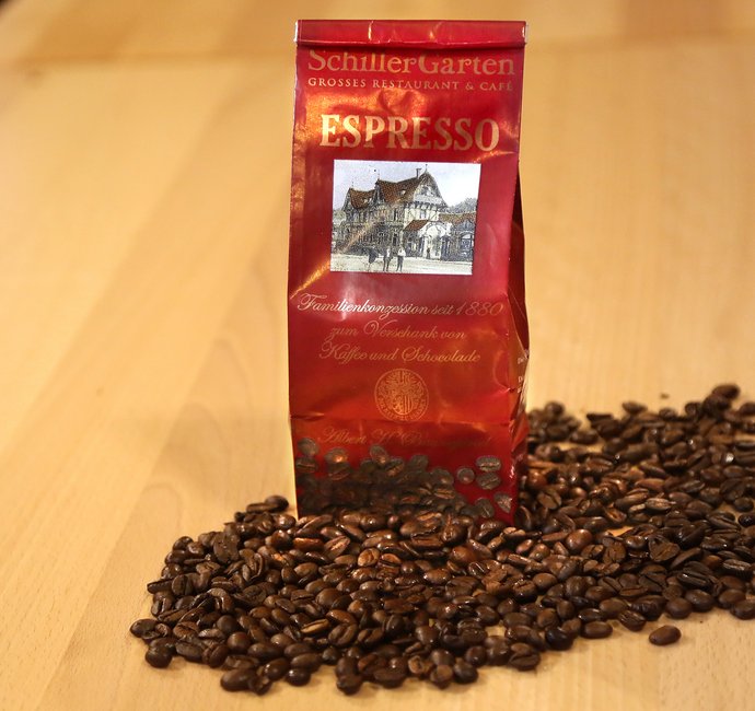 Eine Tüte steht in losen Kaffeebohnen. Auf der Tüte ist der Schriftzug "SchillerGarten. Espresso. Familienkonzession seit 1880 zum Verschank von Kaffee und Schokolade" zu sehen.